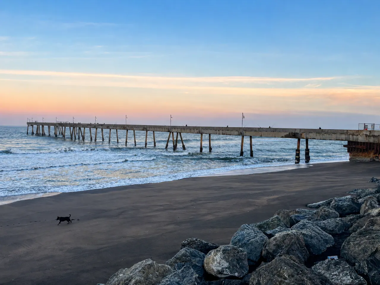 The Pacifica Municipal Pier at dusk with a dog crossing the wet sand and riprap boulders in the foreground