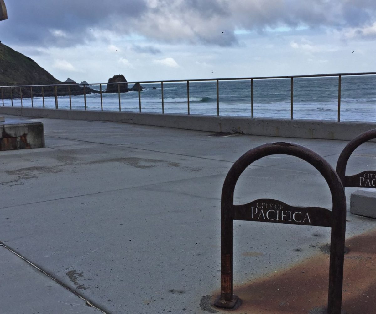View of Rockaway Beach in Pacifica, California, showing the oceanfront promenade with a metal sculpture on the left and two rusted bike racks labeled “City of Pacifica” in the foreground. Waves crash in the distance under a cloudy sky.
