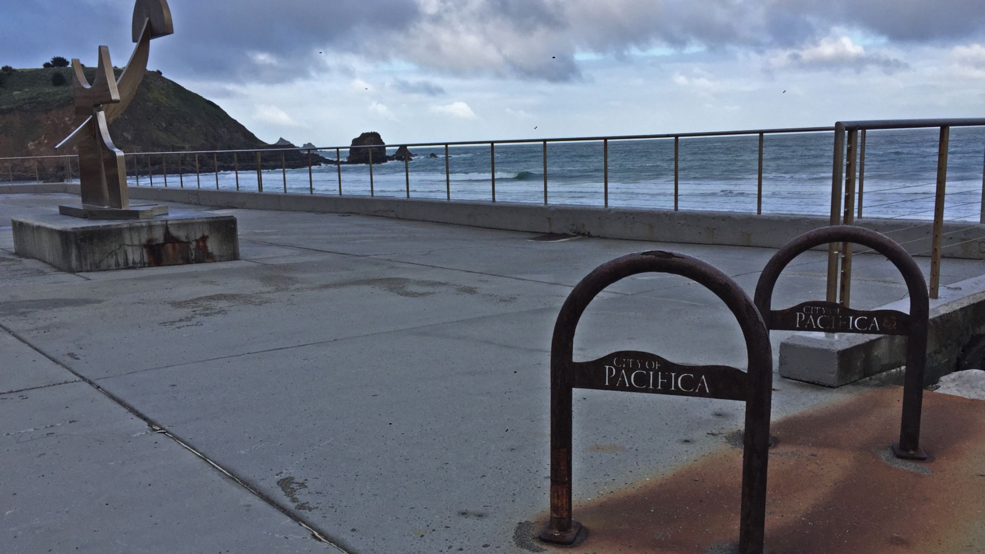 View of Rockaway Beach in Pacifica, California, showing the oceanfront promenade with a metal sculpture on the left and two rusted bike racks labeled “City of Pacifica” in the foreground. Waves crash in the distance under a cloudy sky.